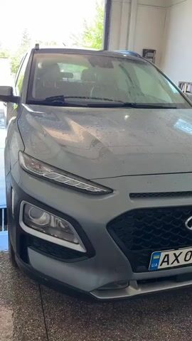 The video shows the front of a gray Hyundai Kona parked indoors, likely at a car wash. The car's hood is covered in water droplets, indicating it is wet, possibly from being washed. The headlights and grille are visible, and a Ukrainian license plate is present. The background is blurred, but appears to be a garage or car wash bay with light coming in from the top.