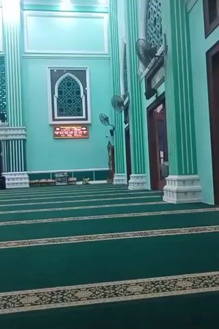 The video pans across the interior of a mosque. The walls and pillars are painted a light turquoise color, with white decorative accents. The floor is covered with rows of green carpet marked with intricate golden patterns. In the background, a large arched niche with decorative patterns is visible, behind which is a window. To the right of this niche, a digital clock or display board shows text in a reddish-orange color. Two ceiling fans are mounted on the wall. Near the entrance on the right, a wooden door is visible. In the foreground, the green carpet stretches out in neat rows. A young person wearing a headscarf is sitting on the carpet in the middle distance. Further back, two young boys, one wearing a cap, walk across the carpeted floor. The overall atmosphere appears calm and quiet.