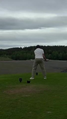 A person wearing a white shirt and tan pants swings a golf club on a golf course. The person is standing on green grass in front of two signs that read "57." The background includes a gray, overcast sky and a landscape with trees and some gray earth. The person swings the club and completes the swing with their arms extended.