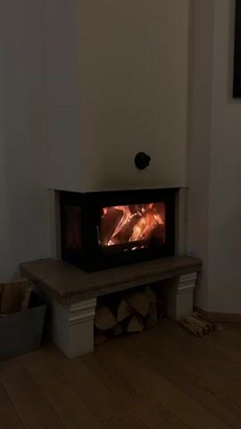 The image captures a cozy indoor scene, likely in a living room, featuring a modern fireplace in a dim setting. The primary focus is a rectangular fireplace with a glass door, inside which a vibrant fire burns, casting warm orange and yellow light. Logs of wood are actively consumed by the flames.

The fireplace is integrated into a white, corner-like structure, with a flat, stone-like hearth or mantelpiece extending outwards. Beneath this hearth, a neat stack of cut firewood is stored, suggesting readiness for use. To the left of the fireplace, a grey bucket or container holds additional smaller pieces of wood, while a few larger pieces rest directly on the wooden floor to the right. A small, black circular knob is visible on the white wall directly above the fireplace opening.

The room is dimly lit, with the fire being the main source of illumination, suggesting it is evening or night. The floor is made of polished wooden planks. The overall ambiance is one of warmth, comfort, and relaxation, characteristic of a chilly evening. The context provided indicates this scene is in Bansko, Bulgaria.