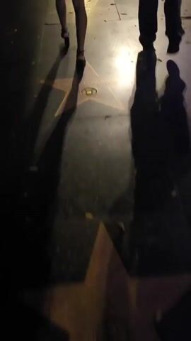 Two people are walking on a sidewalk at night, casting long shadows. They are walking past a series of stars embedded in the sidewalk, which appear to be part of the Hollywood Walk of Fame. The stars are bronze, with a pinkish hue, and have circular emblems in the center. The names on the stars are partially obscured by shadows and the camera's perspective, but visible text includes "CAROL BURNETT". The lighting suggests artificial light sources, such as streetlights or building lights, illuminating the path. The overall mood is dim and atmospheric, with fallen leaves scattered on the ground. The setting is clearly an urban environment, likely a busy street, given the presence of the Walk of Fame.