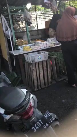 The video shows a market scene with several people, mostly women, browsing at a stall. The stall appears to be selling produce and other goods, displayed on tables and in crates. One woman in a brown hijab and black pants is examining items on the table. Another woman in a light pink hijab and black dress is standing next to her, facing the stall. A third woman in a black hijab and patterned shirt is also present. The ground is unpaved and appears to be dirt or gravel, with some debris scattered around. The overall atmosphere suggests a bustling, everyday market environment.
