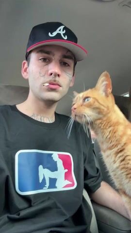 A young man with several face tattoos, a nose ring, and a baseball cap is sitting in the driver's seat of a car, looking at the camera with a cigarette in his mouth. He is wearing a black shirt with a parody MLB logo on it. A light orange cat is sitting on his lap and looking around. The interior of the car is visible.