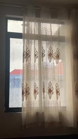 The video shows a window with sheer curtains. The curtains are cream colored with a brown ornate pattern. Sunlight streams through the window, illuminating the curtains. Outside the window, a red roof is visible, suggesting a residential area. The lighting is dim, creating a soft and diffused atmosphere. There are no people or animals in the video, and no discernible actions are taking place. The environment appears to be indoors, likely a room.