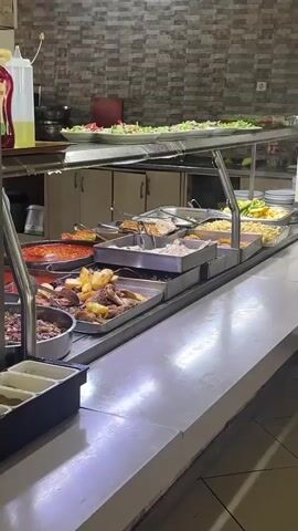 This video shows a buffet-style food service counter in Seydikemer, Turkey. The scene is brightly lit and appears to be indoors, likely a restaurant or cafeteria. The counter is laden with various dishes served in large metal trays and bowls.

In the foreground, to the left, is a stainless steel counter with a few empty compartments. Behind this, a variety of food items are displayed. Prominently featured are trays filled with roasted chicken and potatoes, a large pot of what appears to be a red bean stew or similar dish, and other meat dishes. There are also flatbreads and what looks like a creamy pasta or rice dish. Further back, a salad bar with fresh greens and other salad ingredients is visible on a raised shelf.

A person, likely an employee, can be seen in the background to the right, wearing an apron and tending to the food. They are partially obscured by the buffet setup. The overall impression is of a busy food service environment. The time of day is not discernible, nor is any information about the weather. The setting suggests a casual dining establishment. No visible text is present in the video.