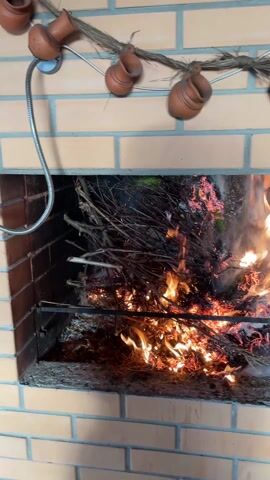 The video shows a brick grill with a fire burning inside. The fire is made up of branches and twigs, and the flames are orange and yellow. There is some smoke coming from the fire. On top of the grill is a decorative rope with small clay pots hanging from it. To the left of the grill is a metal pipe attached to the brick wall.