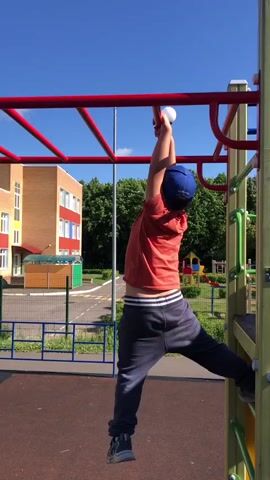 A boy in an orange t-shirt and blue cap is hanging from a red monkey bar on a playground. He is wearing dark pants and dark sneakers. A woman in a blue shirt and jeans stands below him, looking up and reaching out her hand. Another child, a girl in a pink hat and blue shirt, is on the monkey bars to the right of the boy. Behind them is a building, trees, and a blue sky. The ground is covered in red rubber material.