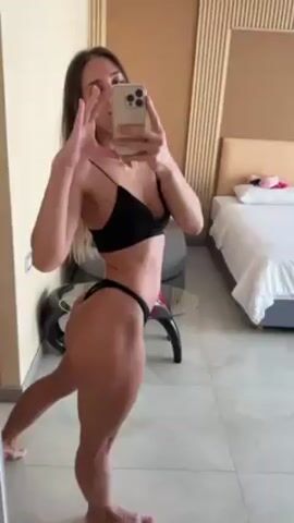 A woman is standing in a room, taking a selfie in the mirror. She is wearing a black bikini. She is holding a phone in her right hand, and her left arm is flexed, showing off her bicep. Her hair is long and brown. The room has a bed, a table, and a tiled floor. There are vertical wooden panels behind the bed. The lighting is bright.