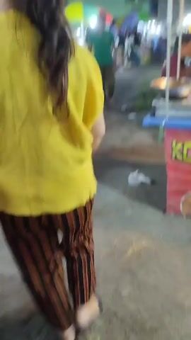 The video shows the back of a person wearing a yellow shirt and striped pants walking past a food stall. The food stall has several trays of food on display. There are other people in the background, and the scene appears to be outdoors at night, possibly a market or street fair. The camera is moving, following the person in the foreground.