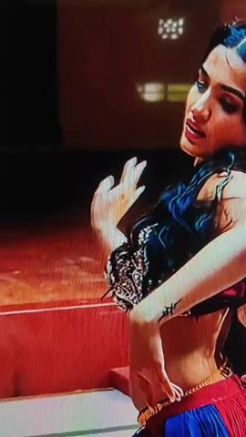 The video begins with a close-up of a woman with long dark hair and intricate jewelry dancing in a dimly lit room. She wears a dark top and a red skirt with blue accents. Her hands are raised and adorned with henna. The camera then pans out to reveal a group of women, all dressed in similar red outfits and performing a synchronized dance. They are on a checkered floor in front of a building with decorative pillars and what appears to be an open courtyard. The lighting is bright, casting shadows on the floor. The dancers move their arms and bodies in unison, creating a dynamic and visually engaging performance.