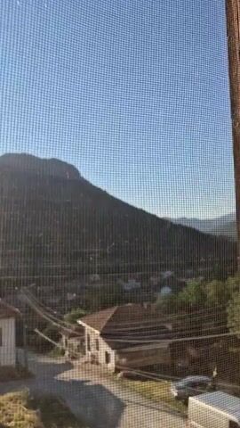 This video shows a view through a window screen of a village in Lesidren, Bulgaria, with a mountain in the background. The sun is rising and casting a warm glow over the scene. The sky is a clear blue, and the weather appears to be pleasant. The village consists of several houses with red-tiled roofs, and there are trees and vegetation scattered throughout. The overall mood is peaceful and serene. The video is filmed from a static position, likely from inside a building. The time of day is early morning, as indicated by the rising sun. There is no visible text in the video.
