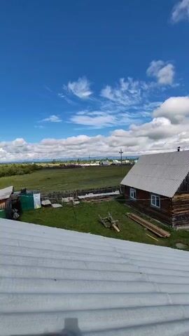 This is a vertical, wide shot of a rural landscape under a blue sky with scattered white clouds. The foreground is dominated by a corrugated metal roof. Beyond the roof, a wooden house with a dark wooden exterior and a lighter-colored roof is visible on the right. Several windows are visible on the house. To the left of the house, there is a fenced area with a green tarp. In the middle ground, there is a grassy field with some scattered wooden planks and a pile of logs. A fence runs along the edge of the field. In the background, there are more houses and structures, suggesting a village. The horizon shows rolling hills and fields. The overall impression is of a peaceful, rural setting on a sunny day.