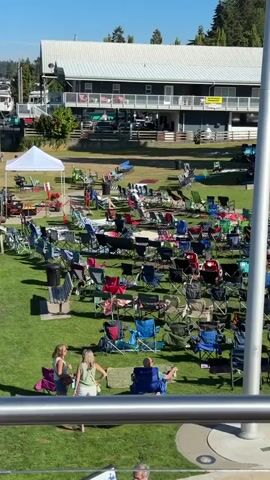 The video opens with a view of a grassy outdoor area filled with numerous portable chairs, many of which are occupied. In the background, there is a building with a white roof and a multi-story structure. Further into the scene, a stage is set up under a covered pavilion, with musical equipment visible. Several tents, some white and some green, are also set up in the area. Two women are standing in the foreground, facing away from the camera, with two people sitting in chairs behind them. The sky is clear and blue, and trees are visible in the background. The overall impression is of an outdoor event, possibly a concert or festival, where people are gathered to watch a performance. The timestamp at the beginning of the video indicates it starts at 00:00. The speaker mentions leaving at approximately 7:00.