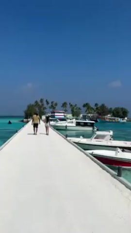 The video shows a man and a woman walking away from the camera on a white pier, towards a small, tree-covered island in the distance. The water on either side of the pier is a clear turquoise color.  Boats are docked on the right side of the pier. On the left side of the pier, there is a jet ski in the water, and further out, a floating inflatable obstacle course with red, white, and blue elements. The sky is clear and blue.
