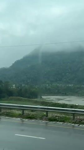 A static eye-level shot shows a road on a gloomy, overcast day. The road is wet, reflecting the gray sky. A silver guardrail runs alongside the road, separating it from a green grassy area, which then leads to a dry riverbed. Beyond the riverbed, a lush green mountain rises, partially obscured by low-hanging clouds. Buildings are visible nestled among the trees on the mountainside. As the video progresses, lampposts appear along the road. A motorcyclist wearing a green rain jacket passes by. The overall impression is one of a rainy, misty day in a mountainous region.