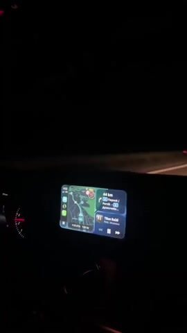 The video, shot from inside a moving vehicle at night, shows the car's dashboard with a digital display indicating navigation and music playback. The road ahead is dimly lit by the car's headlights, revealing a highway with lane markings. The interior of the car is dark, with only the dashboard and the road visible. There are no other cars visible in the immediate vicinity.
