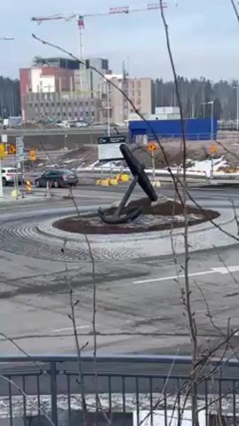 The video shows a roundabout in a city with an anchor statue in the center. Cars are circulating the roundabout. In the background, there is a large building under construction with a crane. Bare tree branches are visible in the foreground. The sky is overcast.