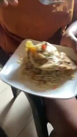 Here is a detailed description of the video:

A person is holding a square white plate with noodles topped with a fried egg. The noodles appear to be light brown and have pieces of red pepper mixed in. The fried egg is large and covers most of the noodles. The yolk appears to be runny and the edges are slightly browned. A fork is used to break apart a piece of the egg. The person is sitting down, with one leg visible. The background includes a green patterned tablecloth and what appears to be a head of lettuce. The lighting is bright, and the focus is primarily on the plate of food.