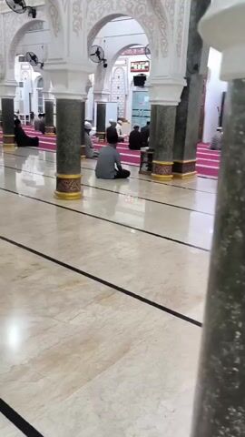 The video shows an interior view of a mosque. The floor is polished marble with black lines. Several people are seated on a pink carpet, facing towards the right of the frame. They appear to be engaged in prayer or religious observance. The mosque features large white pillars with decorative gold accents and arched windows with intricate patterns. Fans are visible on the ceiling. The lighting is bright, creating reflections on the polished floor.