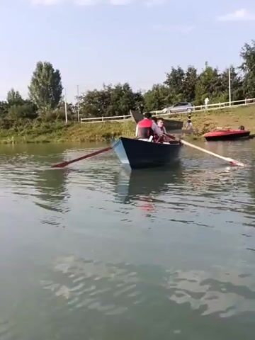A person is sitting in a boat with their legs out, wearing shorts and shoes. Their feet are resting on pedals, indicating they are about to operate the boat. The boat is on a body of water, with trees and a white car visible on the shore in the background. The water is calm and reflects the sky.