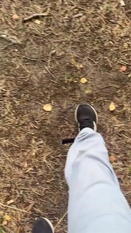 The video shows a first-person perspective of someone walking on a dirt path. The path is covered with leaves and twigs. The person is wearing light-colored pants and dark shoes. As the person walks, the camera pans down to show their feet and then briefly focuses on their hand as they reach out. The background consists of green grass and some trees. The lighting suggests it is daytime.