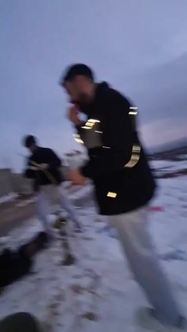 A young person with short brown hair is exhaling a large plume of white smoke. They are wearing a black jacket over a hooded sweatshirt and are outside in what appears to be a cold environment with snow on the ground. The sky is a light blue. In the background, there is a blurry figure standing near a white vehicle. The person holds what looks like a pink vaping device in their mouth.