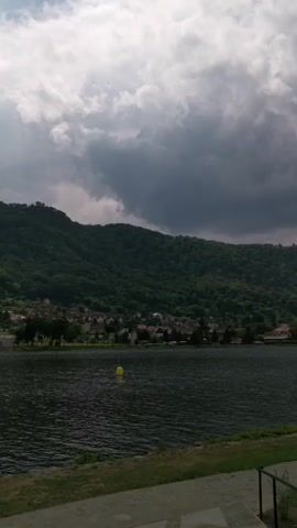 A wide shot captures a body of water with a dark, cloudy sky overhead. A tree-covered hill rises in the background, with a small town nestled at its base. A yellow buoy floats in the water. The foreground shows a stone pathway and grass. The video is still.