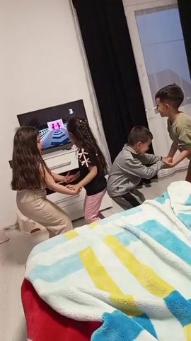 Four children are playing a game in a room with a TV. The children are positioned in a square shape with two children on each side holding hands. They are alternating leaning backwards and forwards, trying to pull each other. On the TV screen, the game shows a dancing character on a brightly colored stage. The TV sits on a white dresser. Behind the dresser, there is a window with black curtains. In the lower right foreground, a bed is covered with a blue, yellow, white, and red blanket. The room has white walls and light-colored wood floors.