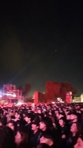 The video captures a night event in Maipú, Chile, showcasing a large crowd of people gathered outdoors. On the right side of the frame, numerous individuals are visible, many looking towards a central stage area. The stage features a metal scaffolding structure illuminated by various lights, including prominent red and white beams cutting through what appears to be smoke or fog effects. The sky is dark, indicating nighttime. The crowd appears engaged, watching the stage, suggesting a concert or festival is taking place. The camera is held vertically and tilted, giving an unusual orientation to the scene. No discernible text is visible.