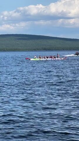 A group of people are in a long boat on a body of water, paddling in unison. The boat appears to be a dragon boat, with a dragon head and tail decoration. The people are dressed in casual clothing. In the background, there is a lush green forest covering a hill, and a blue sky with white clouds. The water is choppy, indicating a gentle breeze. Another inflatable boat is visible to the left of the dragon boat.