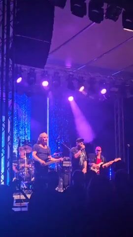 The video shows a live music performance on a stage, illuminated by purple and blue lights and spotlights. There are several band members visible. On the left, a guitarist with long, blonde hair is playing a guitar. Next to him, a singer wearing a cap and a dark shirt is holding a microphone and singing. To the right of the singer, another person in a red shirt and sunglasses is playing a guitar. In the background, a drummer is partially visible behind the singers. The stage is set up with speakers and lighting equipment. In the foreground, the silhouettes of an audience can be seen.
