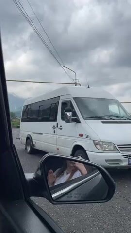 The video captures a view from inside a vehicle, looking out at a street scene. A white minibus is parked to the right of the frame, obscuring some of the background. In the side mirror of the vehicle, a person with blonde hair wearing a white shirt is visible, touching their face. The road appears wet, suggesting recent rain. In the distance, mountains rise under a cloudy sky, and a few buildings and trees are scattered across the landscape. There are utility poles with wires and pipes visible in the middle ground. A sign that says "HOTEL" is partially visible on the right side of the frame. The overall impression is of a slightly overcast day in a rural or semi-rural setting.