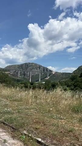 The video shows a long white bridge, supported by tall pillars, spanning across a green valley with a rocky mountain in the background. The sky is blue with scattered white clouds. In the foreground, there is dry grass and some green bushes. The camera zooms in on the bridge, focusing on its structure and the surrounding landscape. There is a short road in the bottom left corner.