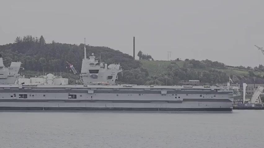 The video captures a large, grey warship, identified by the hull number "R09" visible on its forward island superstructure, slowly moving across a body of water. The ship, which appears to be an aircraft carrier of the Queen Elizabeth-class, is seen from a side perspective, showcasing its expansive flat flight deck and two distinct island superstructures. The camera pans to the left, following the vessel's steady progression.

In the background, a hilly and densely forested coastline dominates the view, indicative of the Norwegian landscape. On the left side of the frame, nearer to the shore, several industrial or port buildings are visible, including a prominent white building marked with the letters "SLB." Other structures, possibly cranes or transmission towers, also dot the distant hills.

The scene is set on an overcast day, with a muted, grey sky contributing to a generally somber lighting. The water surface is calm, reflecting the subdued light. The ship's movement suggests it is either entering or departing a port or navigating a waterway in the Randaberg area of Norway.