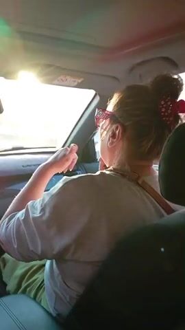 A person with blonde hair pulled up in a bun, wearing pink sunglasses and a light-colored shirt, is seated in the passenger seat of a car. They are seen from the back and side, with their right arm raised and moving in a rhythmic gesture, as if dancing or playing air drums. The sun is shining brightly outside the car, casting a glare on the windshield and dashboard. The interior of the car is visible, including the dashboard and a portion of the driver's seat. The video is a close-up of the person in the passenger seat.