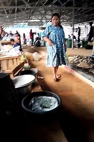 The video shows an indoor market setting. In the foreground, a woman in a blue patterned dress walks past several stalls. To her left, there are large plastic containers. To her right, a stall displays what appears to be food items. In the background, other people are seen browsing and working at various stalls under a covered roof structure. The ground appears to be dirt. The lighting suggests it is daytime. The scene captures a glimpse of daily life and commerce within the market.
