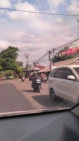 The video shows a street scene in Indonesia, filmed from inside a moving vehicle. The street is lined with small shops and houses, and there are many motorcycles and scooters. The sky is partly cloudy, and the sun is shining. The vehicle is traveling down the street, passing other vehicles and pedestrians. There is a sign for "L.A. Ice Tiga Saudara" visible on the left side of the street. There is a green bus in the distance.
