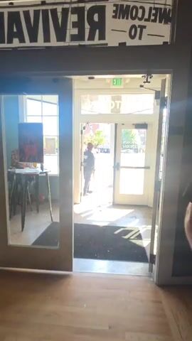 The video captures an interior view looking out through double glass doors onto a bustling city street on a bright, sunny day. Strong sunlight casts distinct shadows of the door frames onto the light-colored wooden floor inside.

A person, dressed in a dark jacket and pants with their hair pulled back, stands in the left doorway, holding what appears to be a strap or rope. They seem to be either entering, exiting, or pausing at the threshold. Outside, a person in a pink shirt and light pants is seen walking past metal barricades on the sidewalk. Other individuals are also visible in the background on the street.

Above the glass doors, a green "EXIT" sign is prominently displayed. The glass transom above the doors reflects "MORTOWN" backwards, suggesting the name "MOTOWN" from an interior perspective. To the right, another glass panel partially shows "BALLROOM & CAFE" in reverse. The street beyond features buildings, trees, and traffic lights, indicating an urban environment.

On the left side of the interior, a small table with a piece of abstract art (red and black) is visible. A black mat lies on the floor just inside the doors. The beginning of the video briefly shows fragmented text like "REVIVAL" and "TO" on a banner overhead.

The audio features a person speaking, mentioning "adversity" and "protesters," which might indicate an event or gathering is taking place inside or nearby. The scene is set in Baltimore, United States of America.