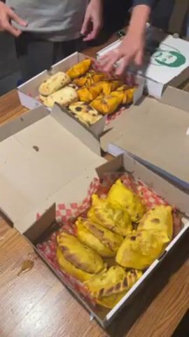 Three boxes are on a wooden table. One box is empty; one contains a dozen yellow pastries on a red and white checkered paper liner; and the third box contains both plain and yellow pastries. The closed box is a pizza box. A man wearing a gray T-shirt and a woman’s hand are visible near the boxes. The boxes contain Bolivian empanadas.