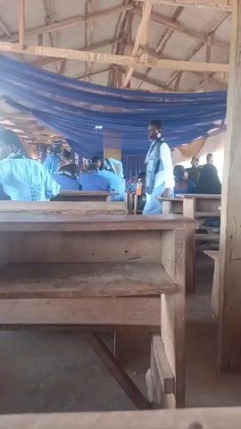 The video shows an indoor event, likely a school function, in Agbara, Nigeria. The setting appears to be a hall with a wooden ceiling and blue drapery. In the foreground, there are traditional wooden school desks.

Several individuals are present, mostly wearing blue attire. One young woman stands out, wearing a white shirt, a light blue tie, and a sash that reads "JUNIOR HIGH" on one side and "ACADEMIC COMMENDATION" on the other, with white fringe. She is holding an orange beverage bottle. Other students are seated at the desks, some facing forward and others turned away from the camera. An adult in a blue cap and gown is also visible, along with other attendees in the background.

The atmosphere seems celebratory, possibly an awards ceremony or a graduation event for junior high students. The sunlight filtering in suggests it is daytime. The overall scene depicts a formal gathering within an educational institution.