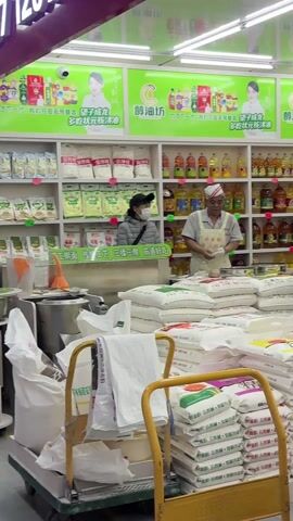 A brightly lit grocery store features a man wearing a white cap and apron standing behind a counter, carefully separating what appears to be dough or batter. He wears white gloves. Behind him, shelves are stocked with various cooking oils and sacks of flour or other dry ingredients. To his left, a woman wearing a face mask and a black cap stands, seemingly waiting. Another person is visible to his right. In the foreground, there's a yellow shopping cart partially visible. Signs advertising products are displayed above the shelves.