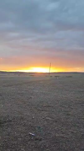 The video shows a wide, open field with sparse vegetation under a cloudy sky at sunset. The sun is setting on the horizon, casting a warm orange glow. There are a few tall poles scattered across the field, likely utility poles. As the video progresses, a chain-link fence with barbed wire appears, leading to a small, utilitarian building with two blue doors. Above the doors, a light fixture is visible. Behind the building, the base of a large lattice tower, possibly a radio or cell tower, can be seen. The overall impression is one of an industrial or communication site in a rural or undeveloped area.