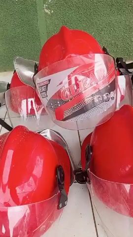 The video shows a close-up, ground-level view of several red motorcycle helmets with clear visors stacked together. The helmets appear to be new or in good condition, with some minor dust or debris visible on the visors and surfaces. Behind the helmets, a pile of red firefighter suits with reflective yellow and silver stripes is visible. The suits are folded or bundled together. The background consists of a green textured surface, possibly a wall or fence, and a tiled floor. The lighting suggests an outdoor or well-lit indoor environment. There are no people visible in the video, and no actions or interactions are taking place. The overall impression is a collection of safety equipment.