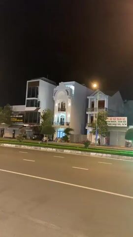 The video shows a row of buildings at night on a street. The buildings are a mix of styles, mostly white, and lit by outdoor lighting. A street runs in front of the buildings with painted lines. A thin strip of green with small plants sits between the street and sidewalk. A streetlight illuminates a portion of the street. Some vehicles pass on the street, and people are seen sitting outside one of the buildings. The sky is dark. Signage in an Asian language is visible on some of the buildings.