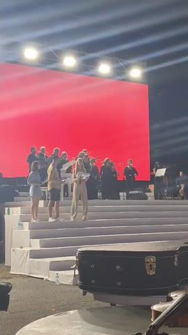 A group of people are on a stage with a large red screen behind them. The screen displays the number "35" and text that reads "GODINA Uz Crnu Goru!". Some people are holding papers and appear to be speaking or singing. Others are positioned behind them, possibly playing musical instruments. In the foreground, a violin case and part of a violin are visible. The stage has white steps leading up to it. The lighting suggests an outdoor or large venue event at night.
