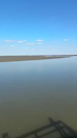 The video shows a wide, calm river or bay under a bright blue sky with scattered white clouds. The water is a murky green-brown color and appears still. On the far bank, a distant shoreline is visible, with what appears to be industrial structures or a city skyline in the haze. In the foreground, a wooden pier or boardwalk is present. The camera pans across the scene, eventually focusing on a blue coin-operated telescope mounted on the pier. The shadow of a railing or structure is visible on the lower part of the frame. There are no people actively shown in the video. The overall impression is one of a tranquil, natural landscape with a hint of human industrial presence in the distance.