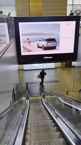 The video shows a downward-moving escalator in what appears to be an airport or shopping mall. Above the escalator, a large digital advertisement display cycles through images: a Range Rover car parked near a body of water with a person standing nearby, followed by a colorful striped pattern, and then a design with geometric shapes. The brand JCDecaux is visible on the bottom of the screen. People can be seen walking in the background on the ground floor. The escalator is made of metal and has glass sides. Yellow lines are painted on the steps, presumably as safety indicators.