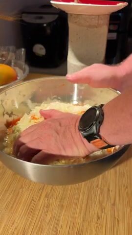 The video shows a close-up shot of a person's hands working with chopped cabbage and carrots in a metal bowl. The person is pressing down on the mixture with both hands, seemingly to pack it or release moisture. The bowl is on a wooden surface. In the background, a black appliance, possibly a rice cooker, and a white object with a red rim are visible. A watch with a black strap and orange accents is worn on the person's right wrist. The lighting suggests an indoor setting, likely a kitchen.
