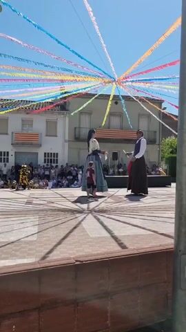 The media file depicts a vibrant outdoor event taking place in a town square. The primary subjects are two tall figures, dressed in traditional, elaborate costumes. The figure on the left wears a long, flowing blue dress with a wide skirt and a dark bodice, and a dark headpiece. The figure on the right is dressed in a white shirt, dark vest, and a long, dark skirt. Between them stands a young boy in a red t-shirt and dark shorts.

The setting is a town square paved with geometric patterns of light and dark tiles, creating a visually striking effect. Behind the figures, a crowd of spectators is gathered, seated and standing, watching the event. The buildings in the background are typical of a European town, with windows and balconies. Two Catalan flags are prominently displayed on the buildings, suggesting the location is in Catalonia, Spain.

Stretching across the sky above the square are numerous colorful streamers, creating a festive canopy of red, yellow, blue, pink, orange, and green. This decorative element, along with the traditional costumes and the gathered crowd, indicates a cultural festival or celebration is underway. The bright sunlight and clear blue sky suggest it is daytime, likely midday, under pleasant weather conditions. The overall atmosphere appears to be one of joy and community engagement. No discernible text is visible in the media file.