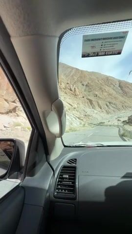 The video captures a first-person perspective from inside a vehicle, looking out through the windshield. The vehicle is driving on a paved road that winds through a mountainous, arid landscape. The mountains are steep and rocky, with sparse vegetation. A river or stream flows alongside the road in some sections, appearing muddy and turbulent. Telephone poles and power lines are visible along the route. The sky is partly cloudy, with the sun casting shadows. A sticker is affixed to the upper right corner of the windshield, appearing to be for emergency roadside assistance. The interior of the vehicle is partially visible, including the dashboard, air vents, and a grab handle. The overall impression is a journey through a remote and rugged terrain.